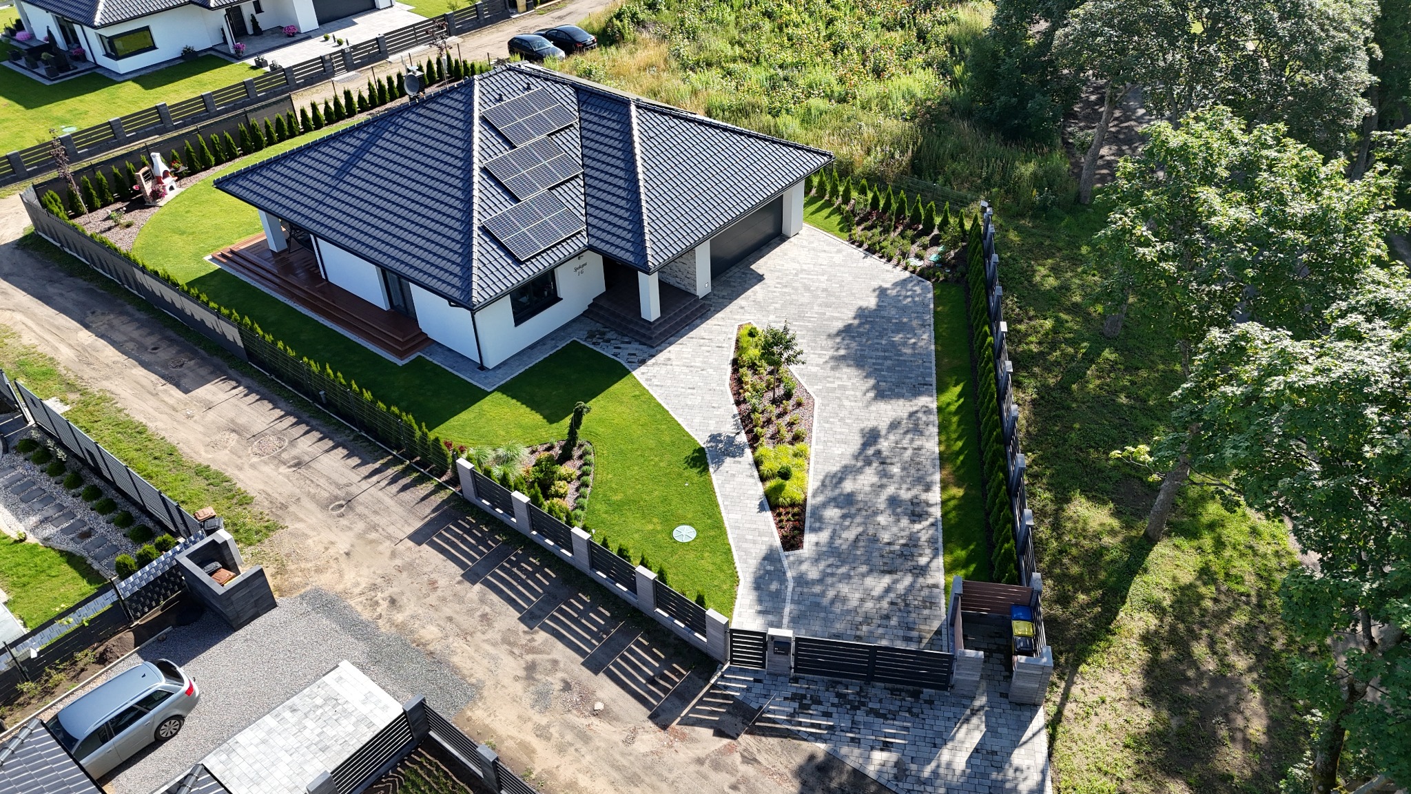 Widok z góry na nowoczesny dom z panelami słonecznymi, zadbanym trawnikiem, ścieżką z kostki i ozdobnymi nasadzeniami wzdłuż ogrodzenia.