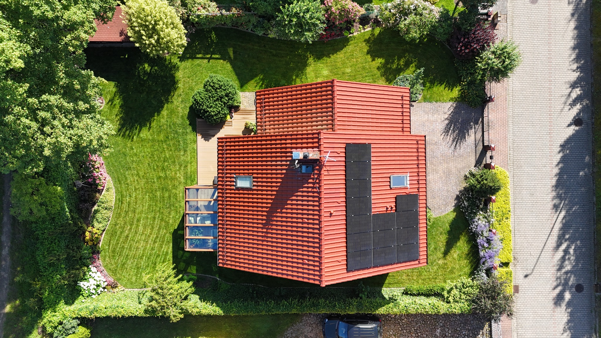 Widok z góry na dom z czerwoną dachówką, otoczony bujnym ogrodem z trawnikiem, drzewami i kwiatami. Panele słoneczne na dachu. Uwagę przykuwa precyzyjnie utrzymana zieleń.