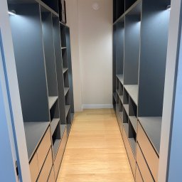 WOBO studio Wojciech Borawski - Wnętrze garderoby z szarymi szafami wnękowymi z oświetleniem LED, jasnymi półkami i szufladami, parkiet na podłodze, widok z perspektywy wejścia.