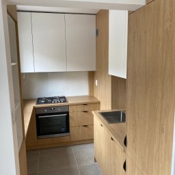 WOBO studio Wojciech Borawski - Jasna kuchnia z drewnianymi szafkami i białymi górnymi frontami. Ustawna zabudowa z płytą grzewczą i piekarnikiem, widok na zlewozmywak. Szare płytki na podłodze.