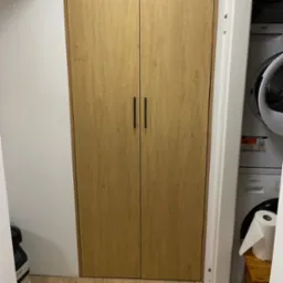 Drewniana szafa wnękowa z czarnymi uchwytami, umieszczona obok pralki i suszarki w pomieszczeniu. Naturalne oświetlenie podkreśla strukturę drewna.