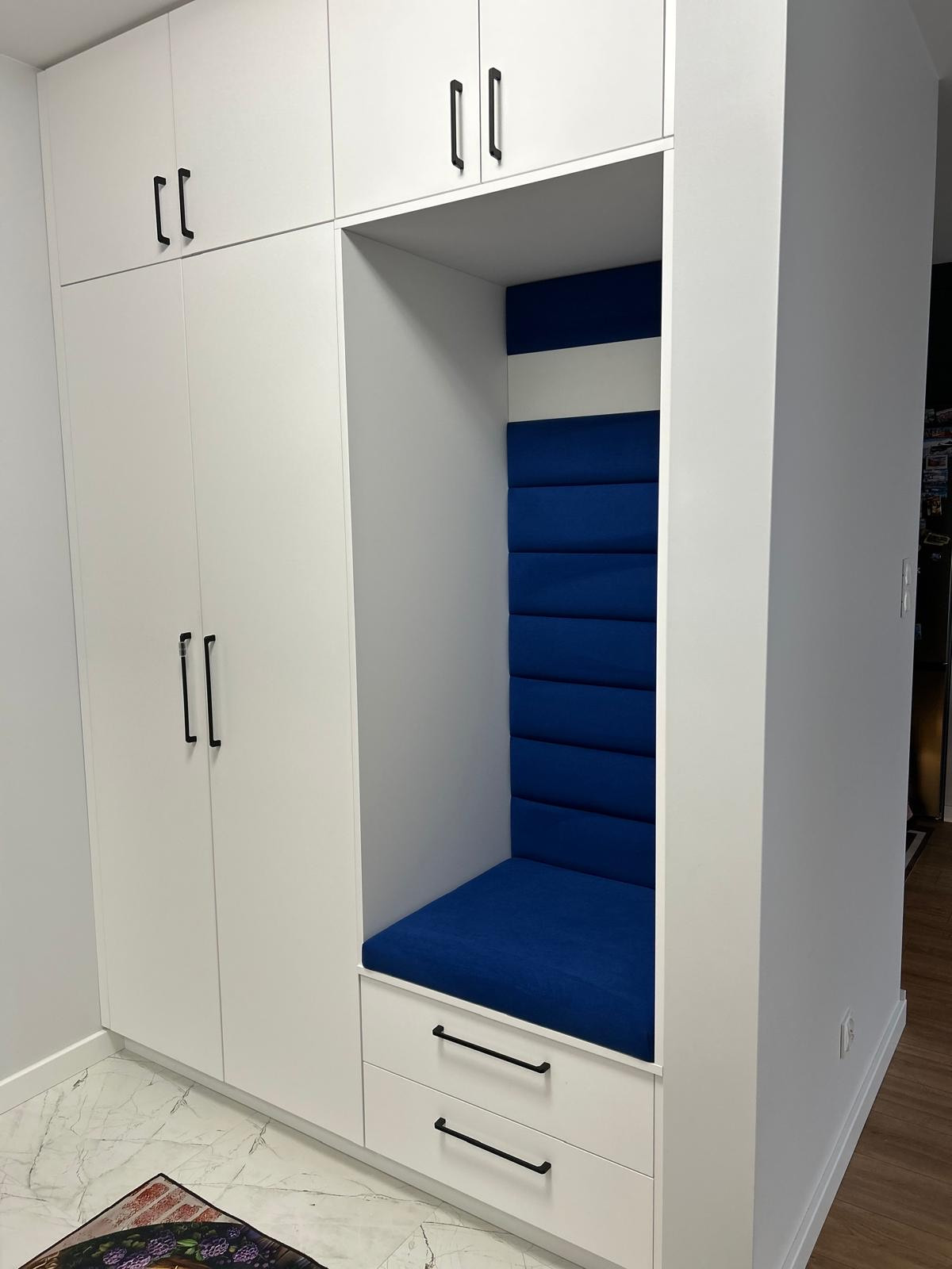 Biała szafa wnękowa z siedziskiem obitym niebieskim materiałem, czarne uchwyty, marmurowa podłoga. Nowoczesny design, zabudowa na wymiar.