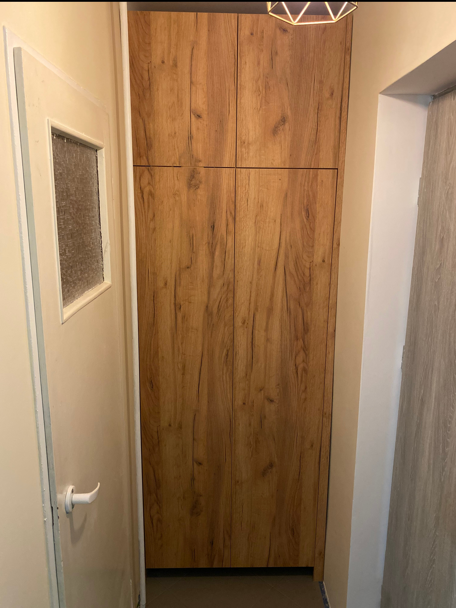 Wysoka, drewnopodobna szafa wnękowa z czterema drzwiami, umieszczona w wąskim przedpokoju obok białych drzwi z szybką.