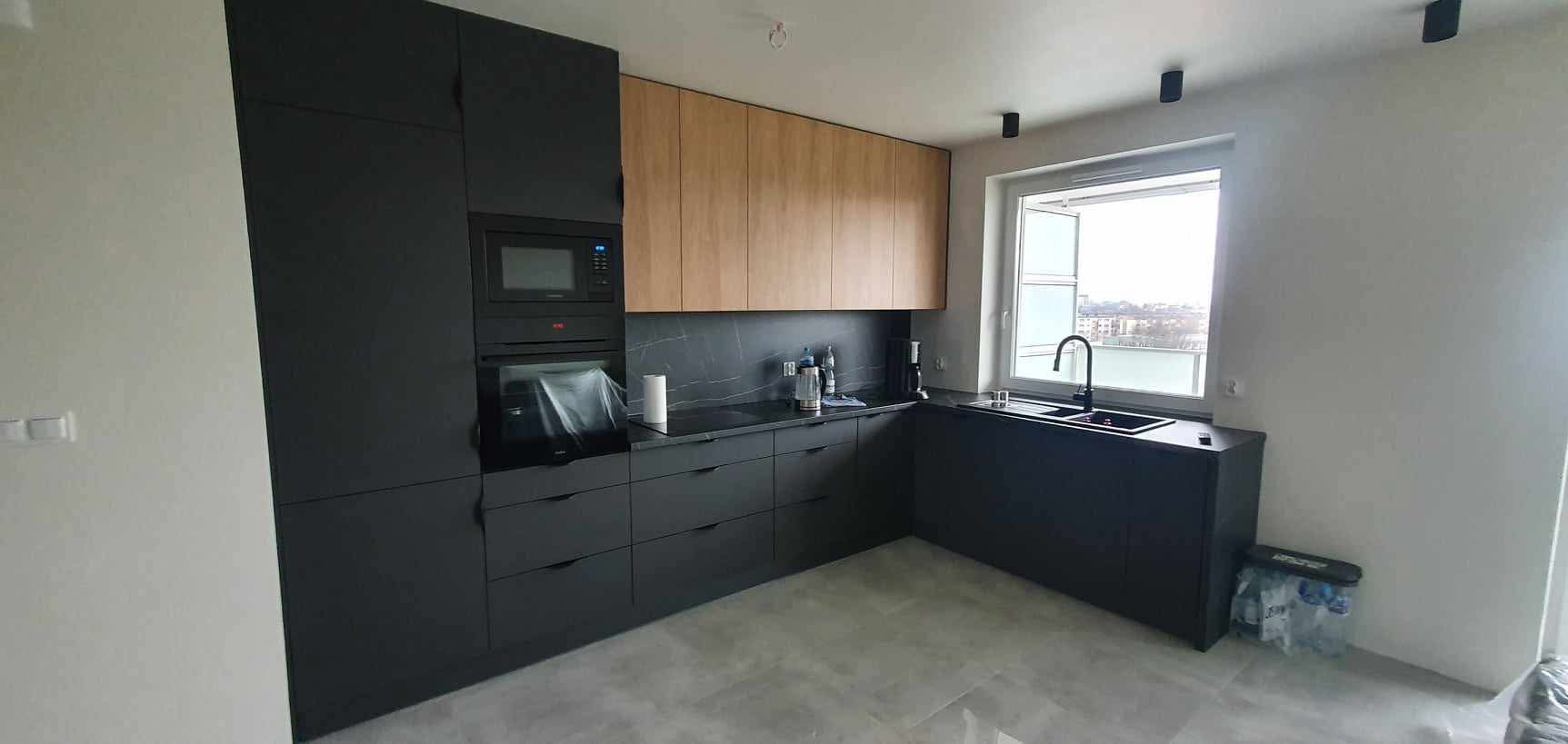 Nowoczesna kuchnia z czarnymi szafkami dolnymi i drewnianymi górnymi, czarny blat i okno z widokiem na miasto. Minimalistyczny design i funkcjonalność.