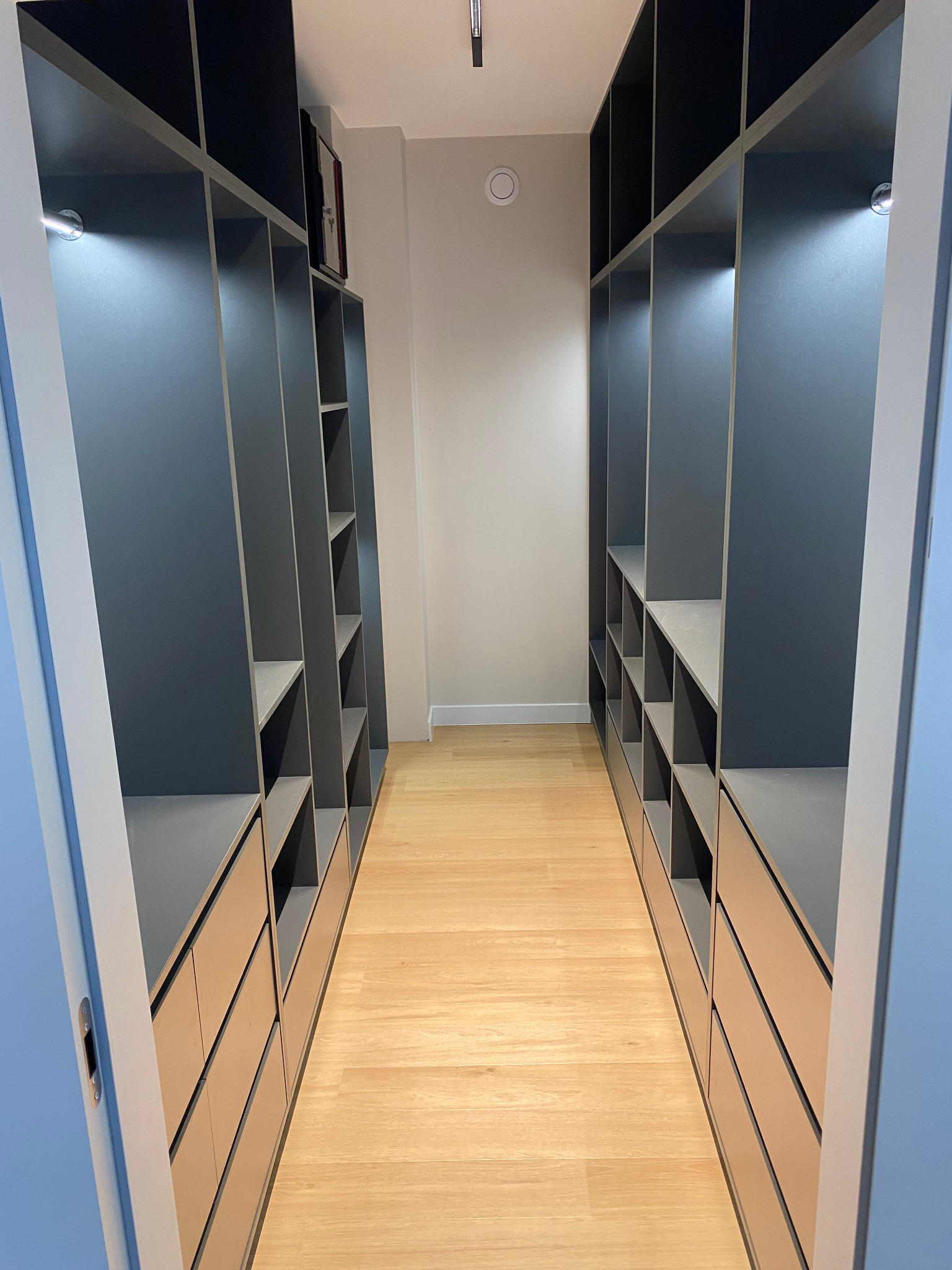 Wnętrze garderoby z szarymi szafami wnękowymi z oświetleniem LED, jasnymi półkami i szufladami, parkiet na podłodze, widok z perspektywy wejścia.