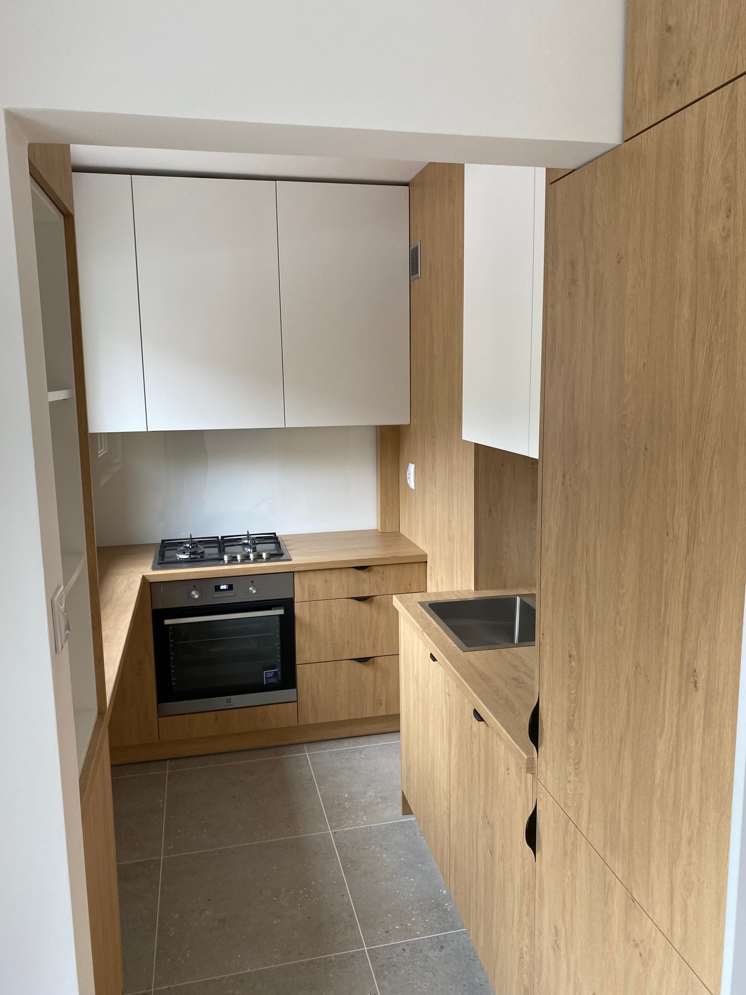 Jasna kuchnia z drewnianymi szafkami i białymi górnymi frontami. Ustawna zabudowa z płytą grzewczą i piekarnikiem, widok na zlewozmywak. Szare płytki na podłodze.