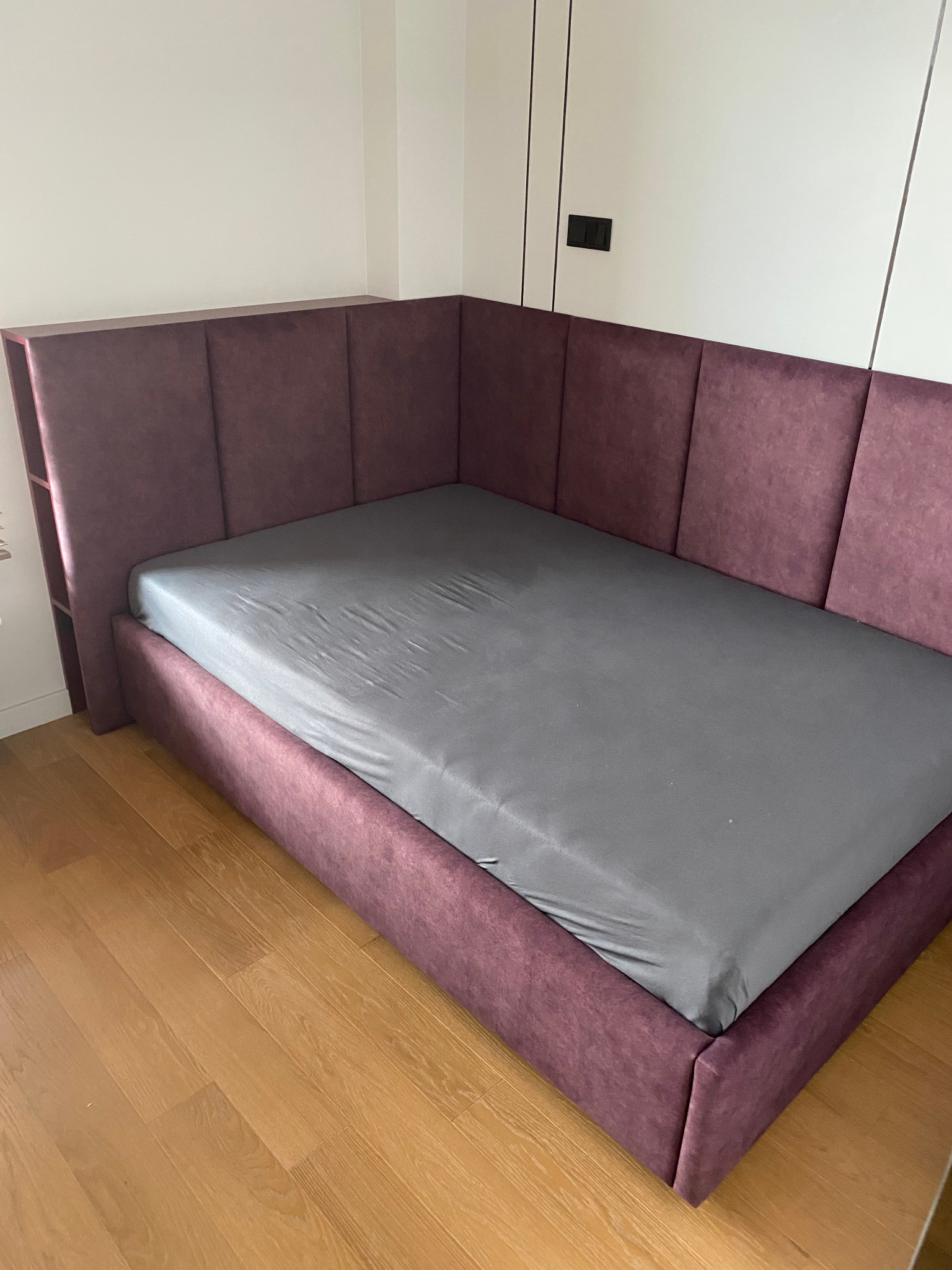 Tapicerowane łóżko w odcieniach fioletu z szarą pościelą, dopasowane do rogu pokoju, z zagłówkiem z półkami. Widoczna drewniana podłoga i białe ściany.
