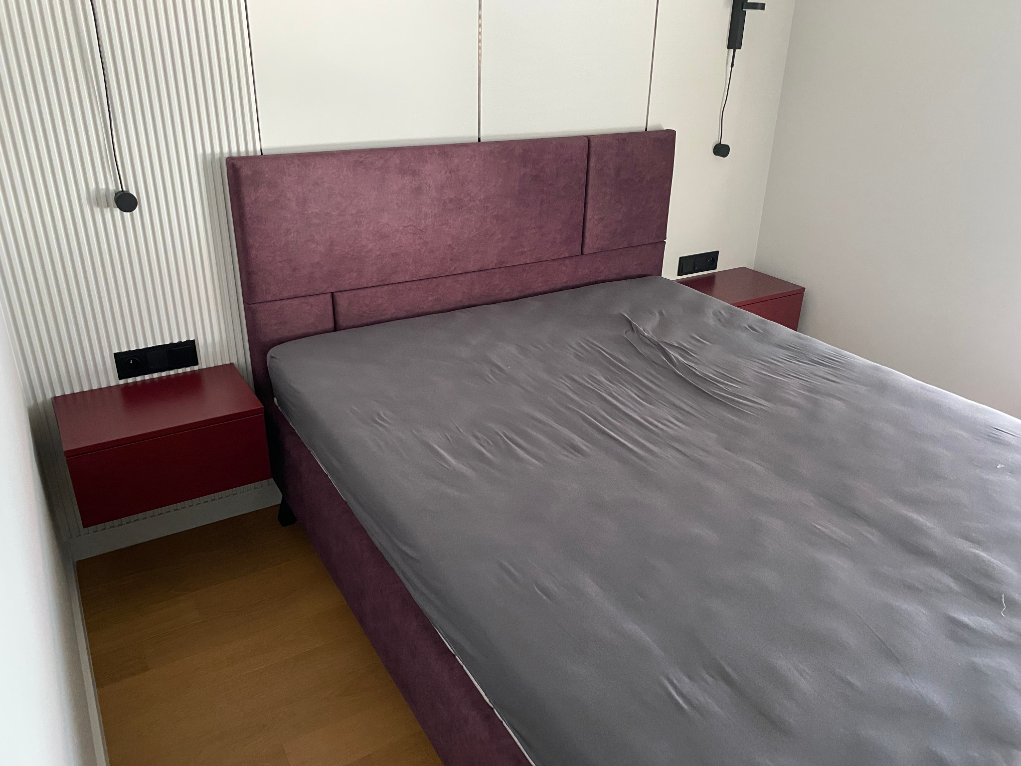 Fioletowe łóżko z tapicerowanym wezgłowiem, z szarym prześcieradłem, w nowoczesnej sypialni z drewnianą podłogą i szafkami nocnymi w kolorze czerwonego wina.