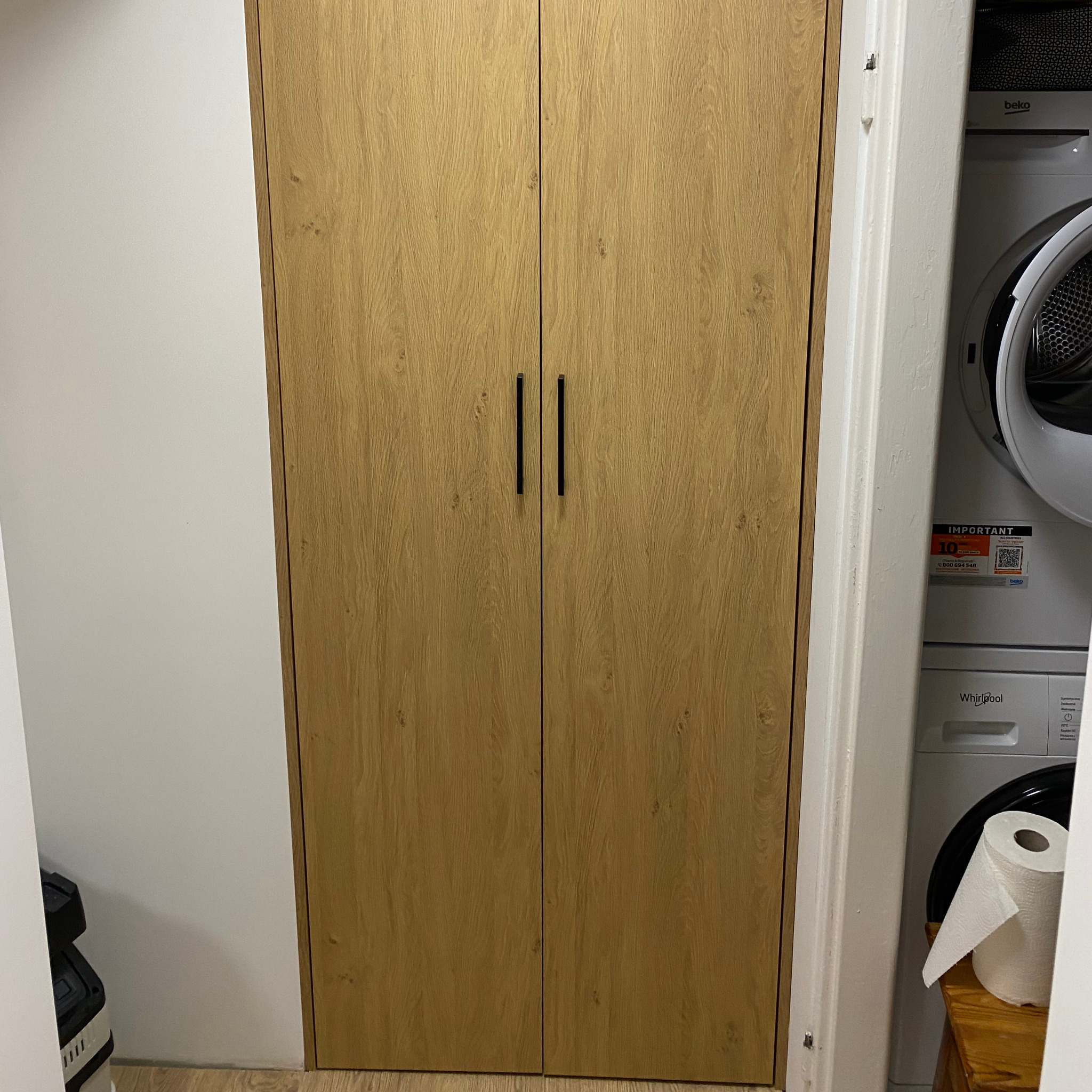 Drewniana szafa wnękowa z czarnymi uchwytami, umieszczona obok pralki i suszarki w pomieszczeniu. Naturalne oświetlenie podkreśla strukturę drewna.