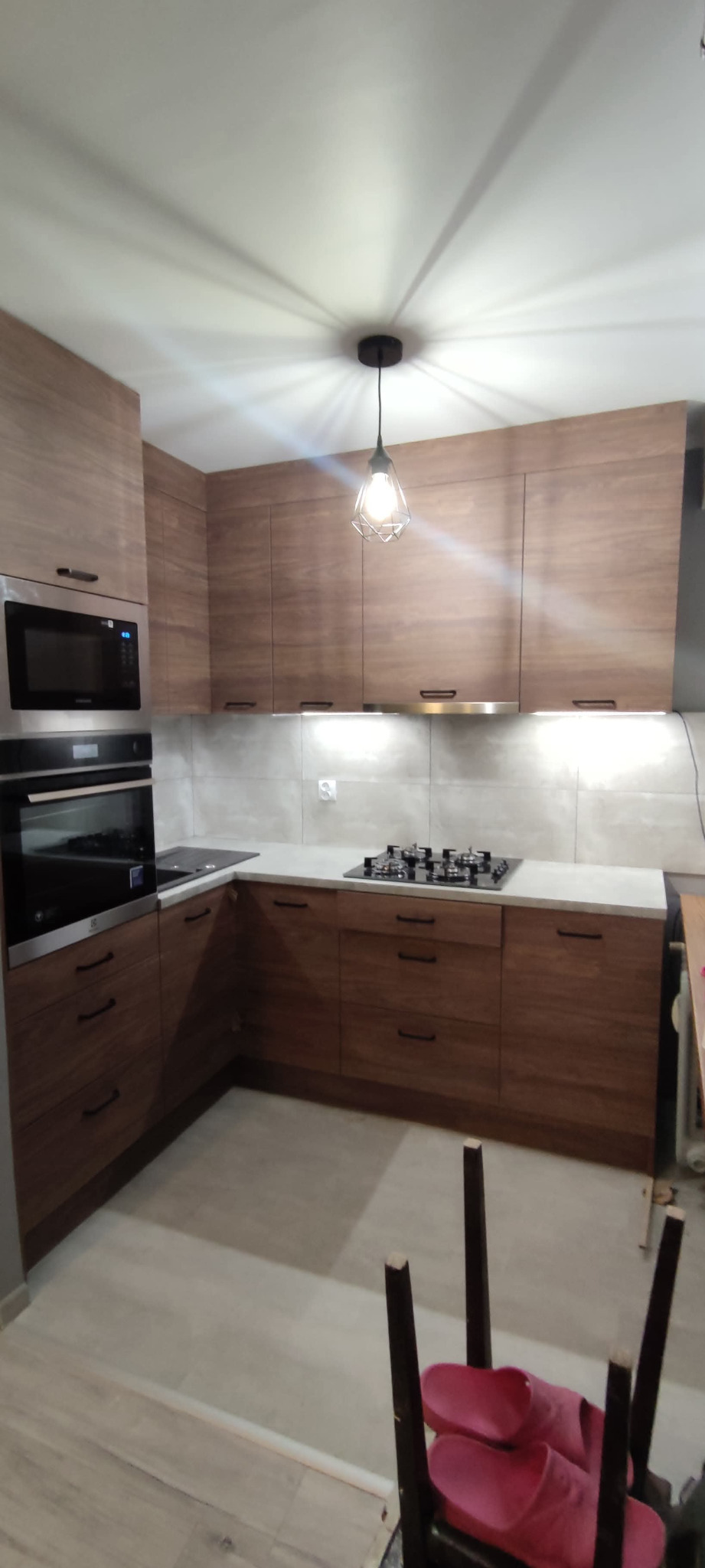 Nowoczesna kuchnia z drewnianymi frontami, białym blatem i oświetleniem podszafkowym. Widoczny piekarnik, mikrofalówka i płyta gazowa. Wisząca lampa dodaje uroku.