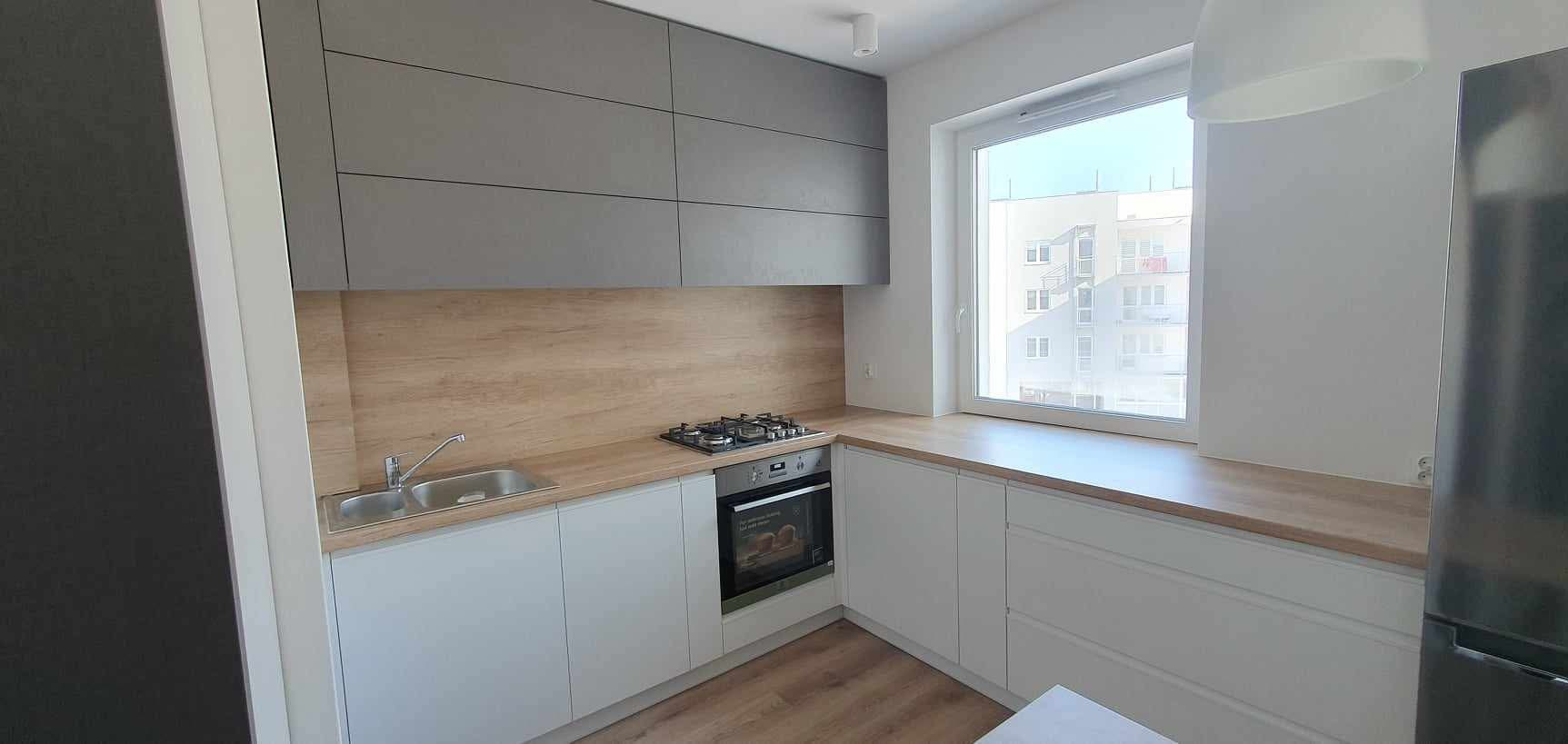 Jasna kuchnia z szarymi szafkami wiszącymi i drewnianym blatem. Widok na zewnątrz przez okno. Nowoczesny design z minimalistycznym stylem.