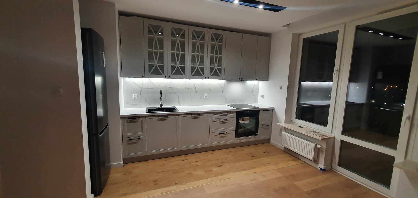 Jasnoszara kuchnia na wymiar z białym blatem i marmurowym panelem. Górne szafki z geometrycznymi przeszkleniami. Widok na okno i podłogę z paneli.