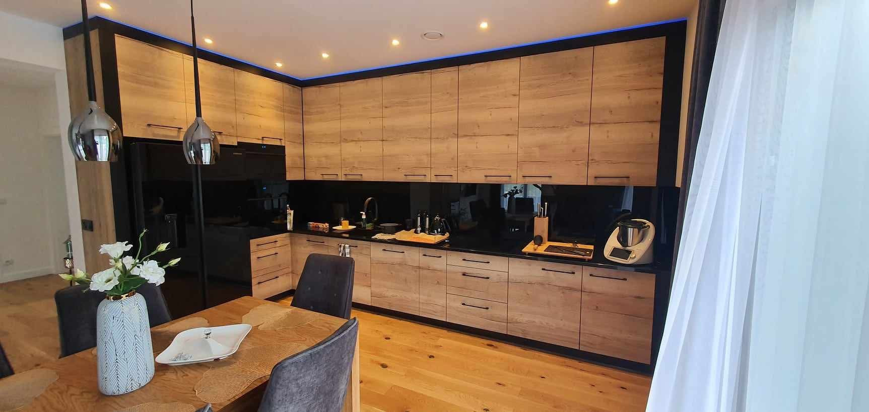 Kuchnia z drewnianymi frontami i czarnym lacobelem, oświetleniem LED i drewnianą podłogą. Na blacie akcesoria kuchenne i Thermomix. W tle stół z kwiatami.