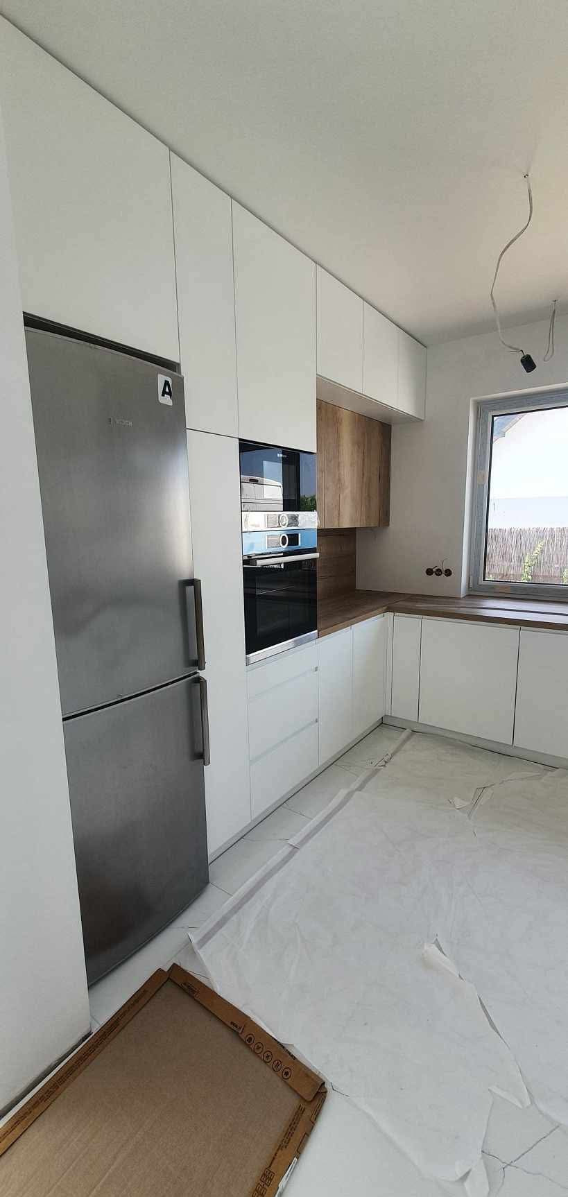 Nowoczesna kuchnia z białymi szafkami, drewnianym blatem i srebrną lodówką Bosch. Podłoga zabezpieczona folią malarską. Widoczne gniazdka i okno.