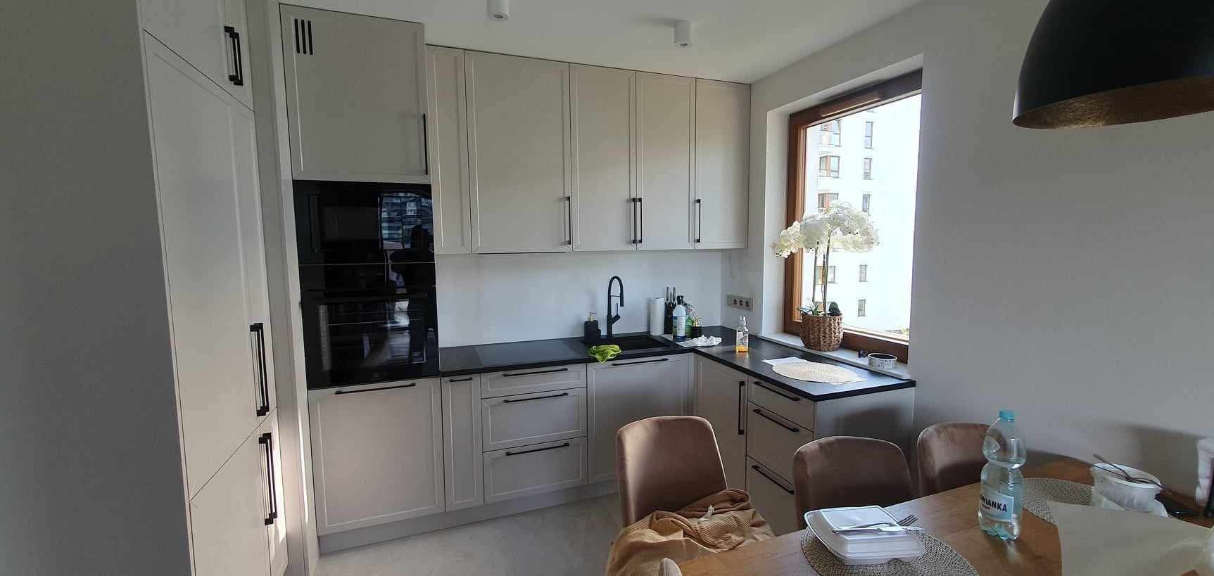 Jasna kuchnia z czarnymi akcentami: zabudowa meblowa z piekarnikiem, szafki wiszące, czarny blat i zlew. Widok na stół z krzesłami i okno z kwiatem.