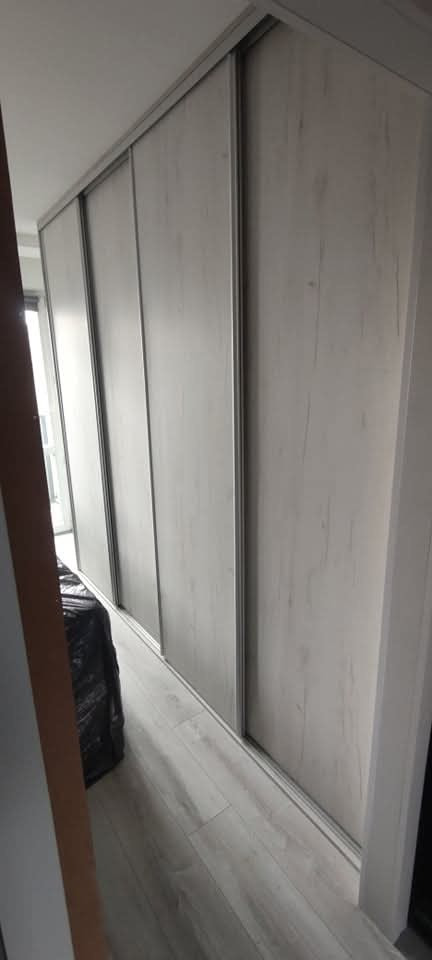 Jasna szafa wnękowa z przesuwnymi drzwiami, imitacja drewna, zamontowana w Białymstoku. Minimalistyczny design, widoczne prowadnice i podłoga w tym samym odcieniu.