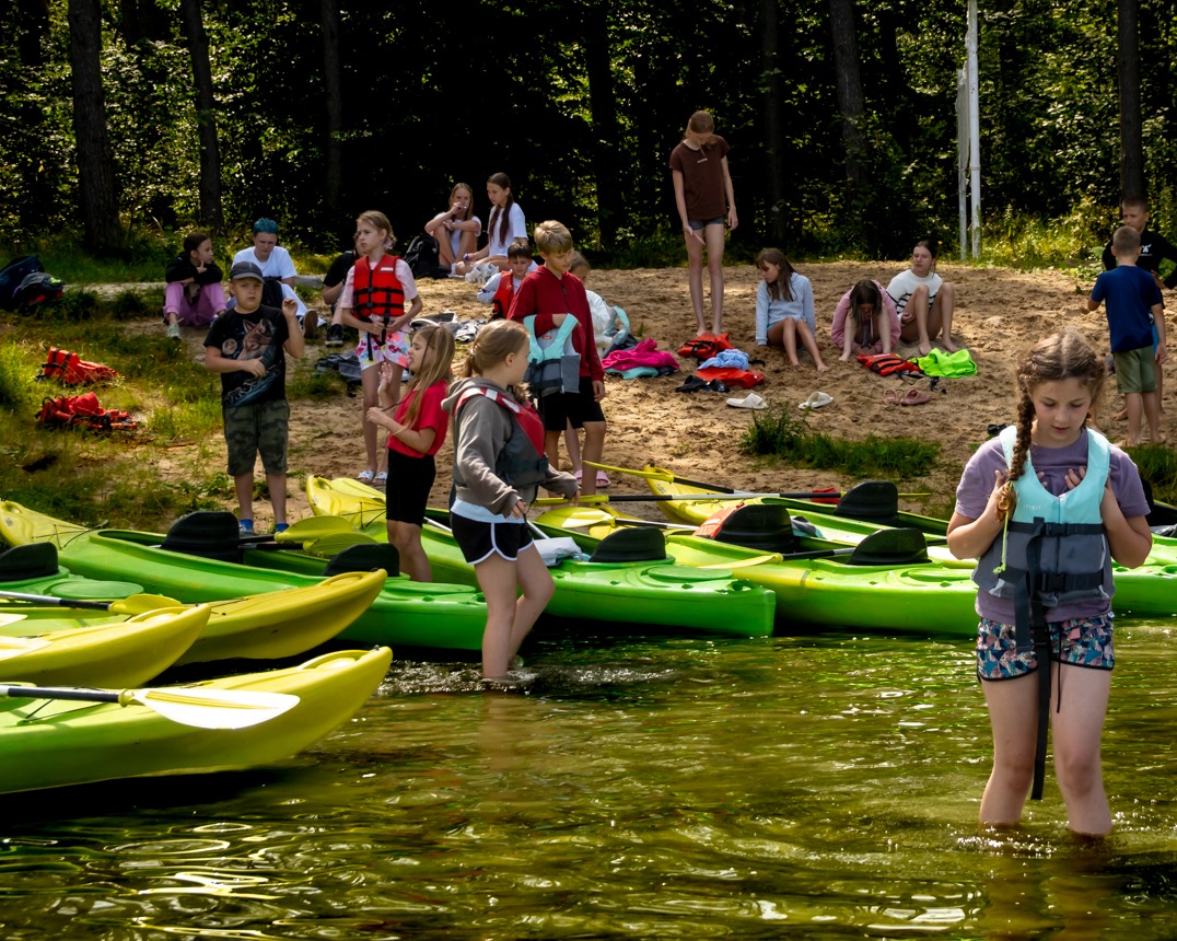 Grupa dzieci w kamizelkach asekuracyjnych przygotowuje się do spływu kajakowego w Grudziądzu. Kajaki na pierwszym planie, w tle las.