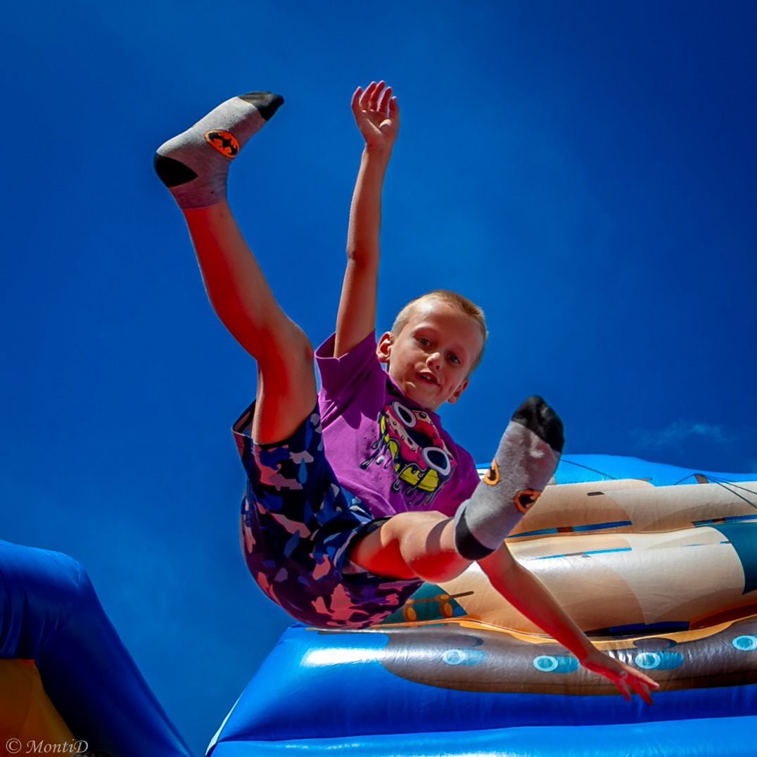 Chłopiec w locie na dmuchanym zamku, ubrany w fioletową koszulkę i skarpetki z logo Batmana, na tle intensywnego błękitu nieba. Dynamiczne ujęcie z dołu.