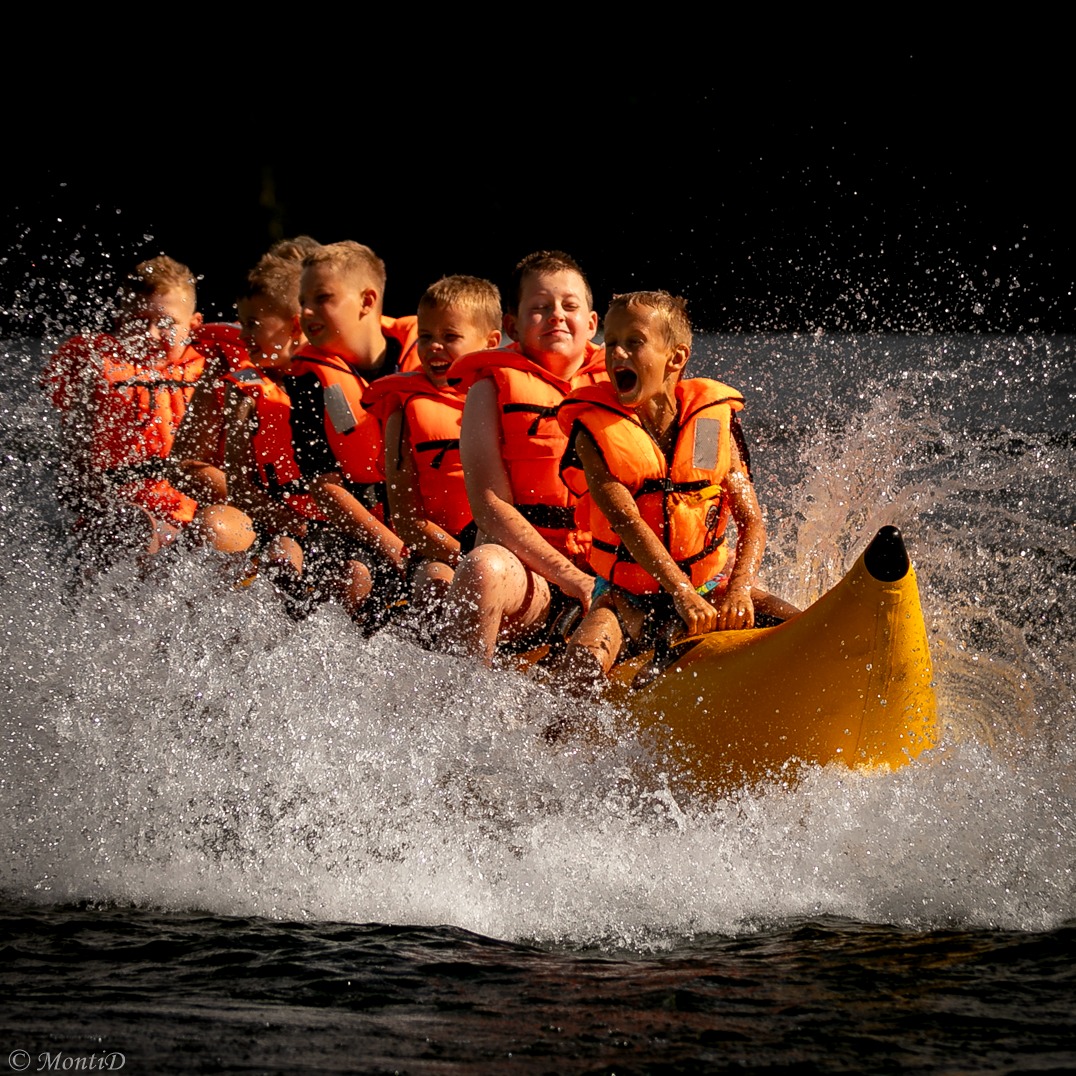 Grupa dzieci w pomarańczowych kamizelkach płynie na dmuchanym bananie po wodzie, otoczona bryzgami. Jeden z chłopców krzyczy radośnie.