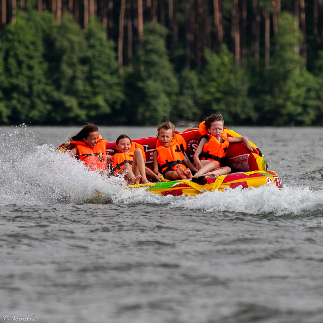 Radosne dzieci w pomarańczowych kamizelkach unoszą się na dmuchanym kole za motorówką, wzbijając fontannę wody. Letnia zabawa na jeziorze w otoczeniu zieleni.