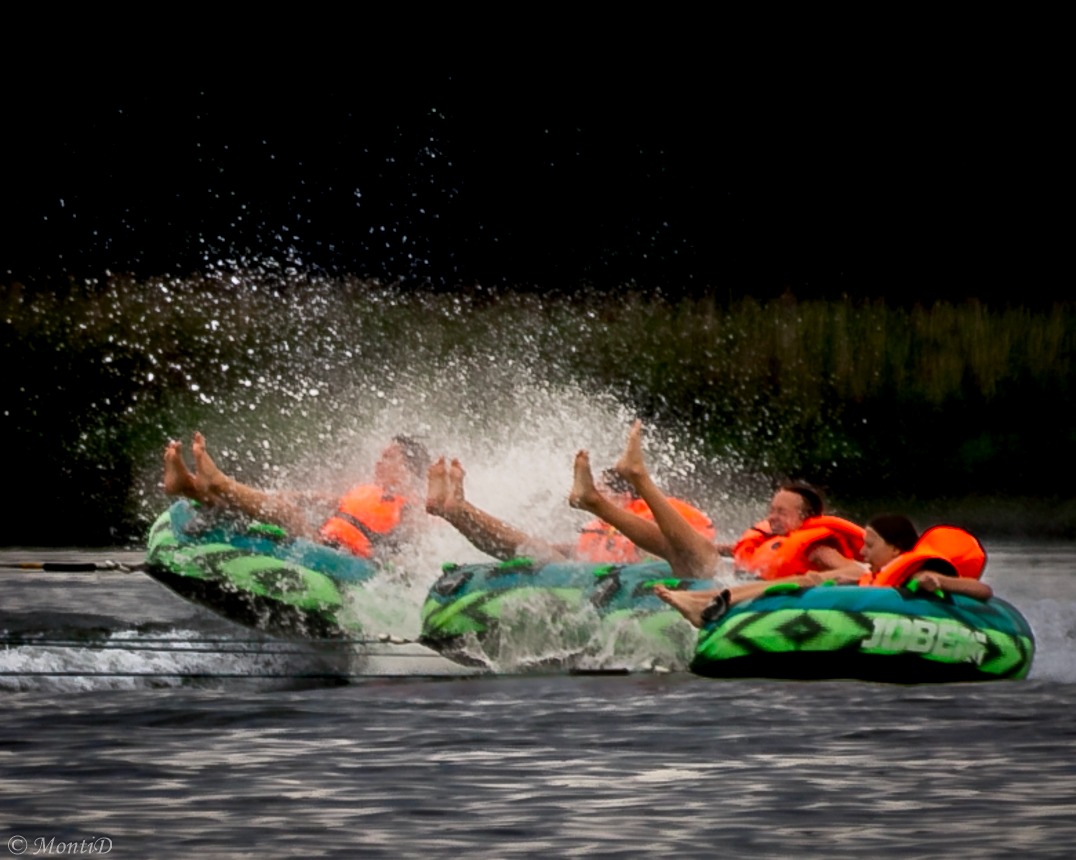 Radosna jazda na dmuchanych kołach po wodzie. Grupa dzieci w pomarańczowych kamizelkach, ciągniętych liną, otoczona bryzgami wody. Aktywny wypoczynek.