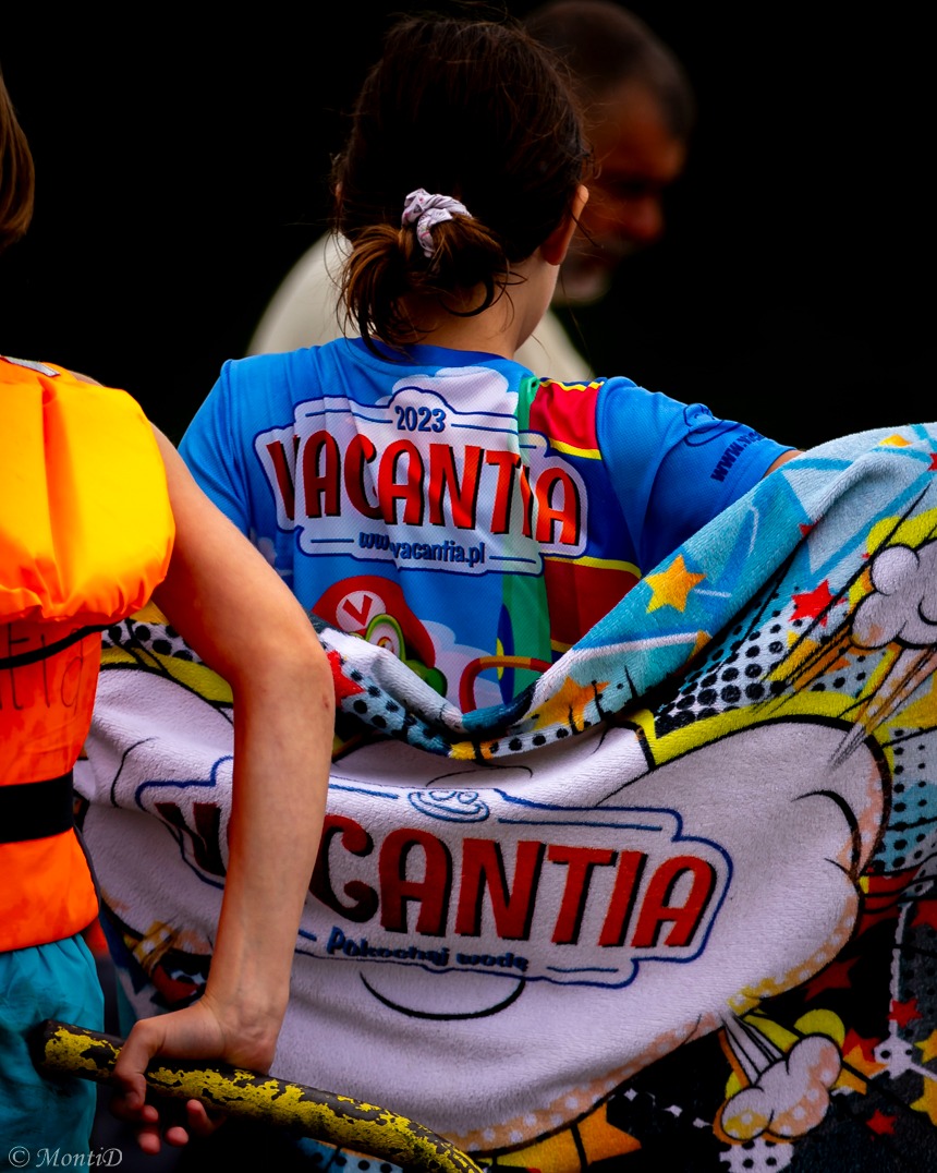 Uczestniczka obozu z ręcznikiem i koszulką Vacantia, widoczna z tyłu, z fragmentem kamizelki ratunkowej, na tle rozmytej postaci dorosłego.