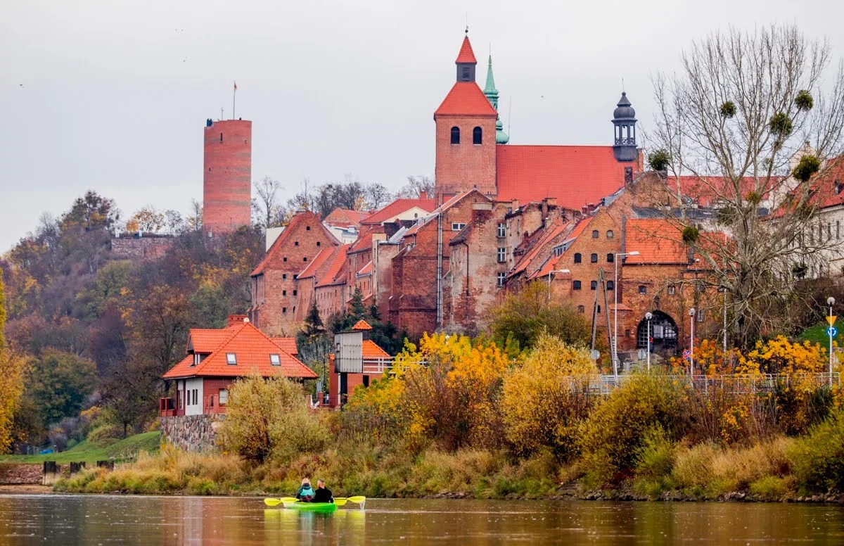 Kajak z dwiema osobami na rzece, w tle ceglane budynki i wieże w Grudziądzu, jesienna sceneria, pochmurne niebo.