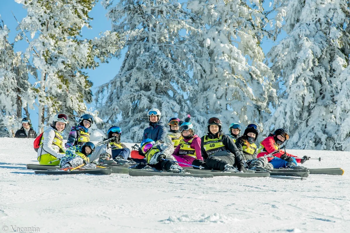 Grupa dzieci w kaskach i goglach siedzi na nartach na śniegu, w tle ośnieżone drzewa i błękitne niebo. Zimowy krajobraz, obóz narciarski.