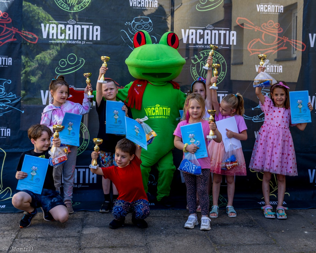 Grupa dzieci z nagrodami i dyplomami na tle baneru Vacantia, z maskotką żabą. Uśmiechnięte twarze, trofea, radosna atmosfera po zakończonych zajęciach.