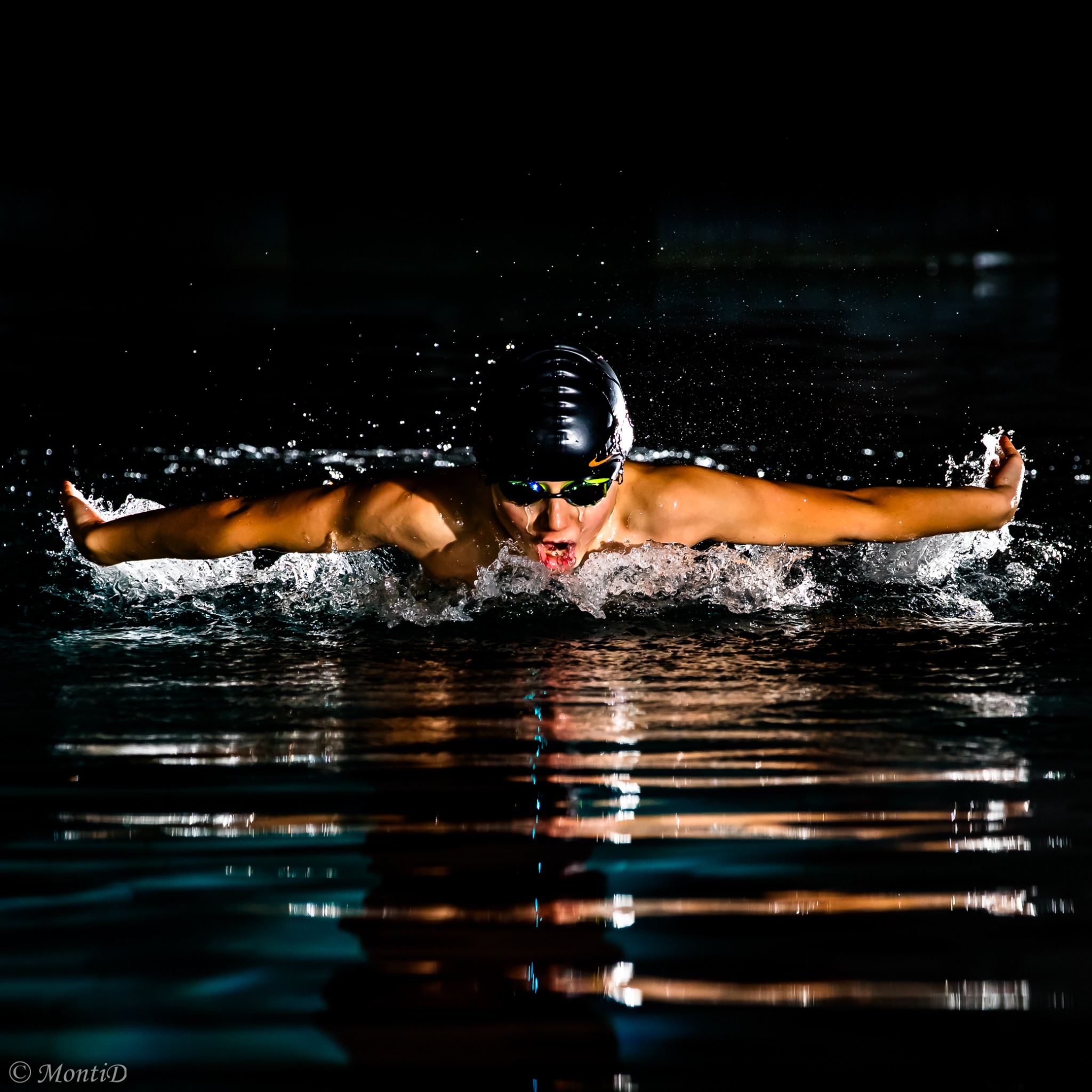 Pływak w czepku i okularach płynie stylem motylkowym w basenie, widoczne odbicie w wodzie i dynamiczne rozpryski wody. Oświetlenie punktowe podkreśla akcję.