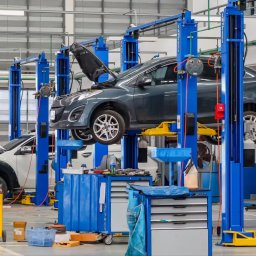 eleasingi.pl LEASING I POŻYCZKA I NAJEM - Szary samochód na podnośniku w warsztacie, z otwartą maską. W tle inne podnośniki i narzędzia. Oświetlenie sufitowe, posadzka betonowa.