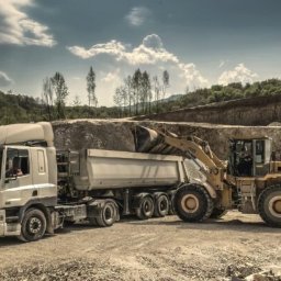 eleasingi.pl LEASING I POŻYCZKA I NAJEM - Żółta ładowarka kołowa ładuje urobek do naczepy ciężarówki na tle kamieniołomu i zieleni. Scena z placu budowy z maszynami w akcji.