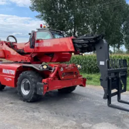 Czerwony teleskopowy wózek widłowy Manitou MRT 2150+ na drodze, z polem kukurydzy w tle. Widoczne detale konstrukcyjne i mocne opony.