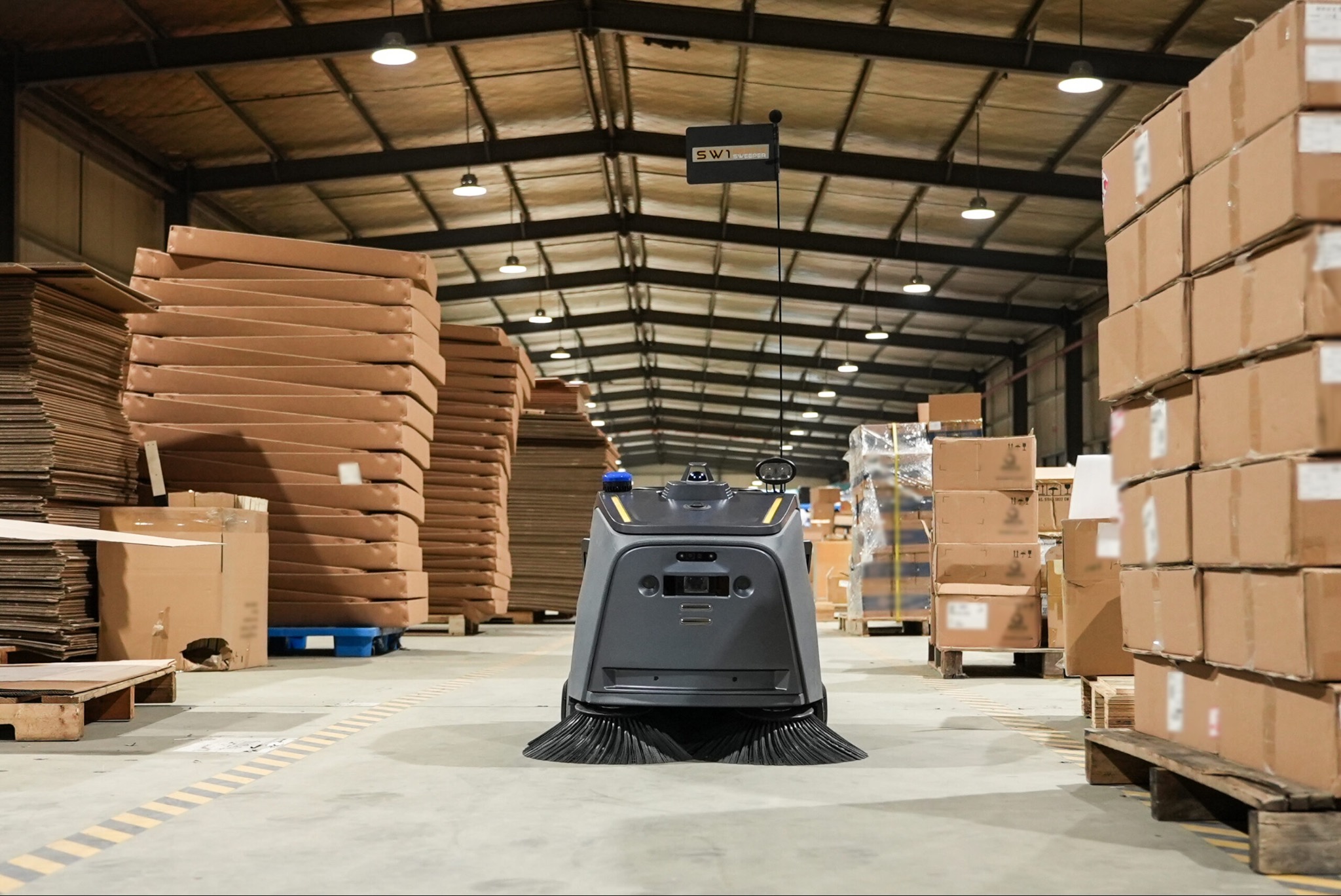 Samojezdna maszyna czyszcząca SW1 Sweeper w magazynie w Gomunicach. Urządzenie do sprzątania hal, z kartonami i oświetleniem w tle. Perspektywa z dołu.