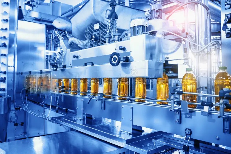 Automatyczna linia produkcyjna: szklane butelki z płynem przesuwają się na taśmie, oświetlone jasnym światłem. Metalowe elementy maszyny w odcieniach niebieskiego.