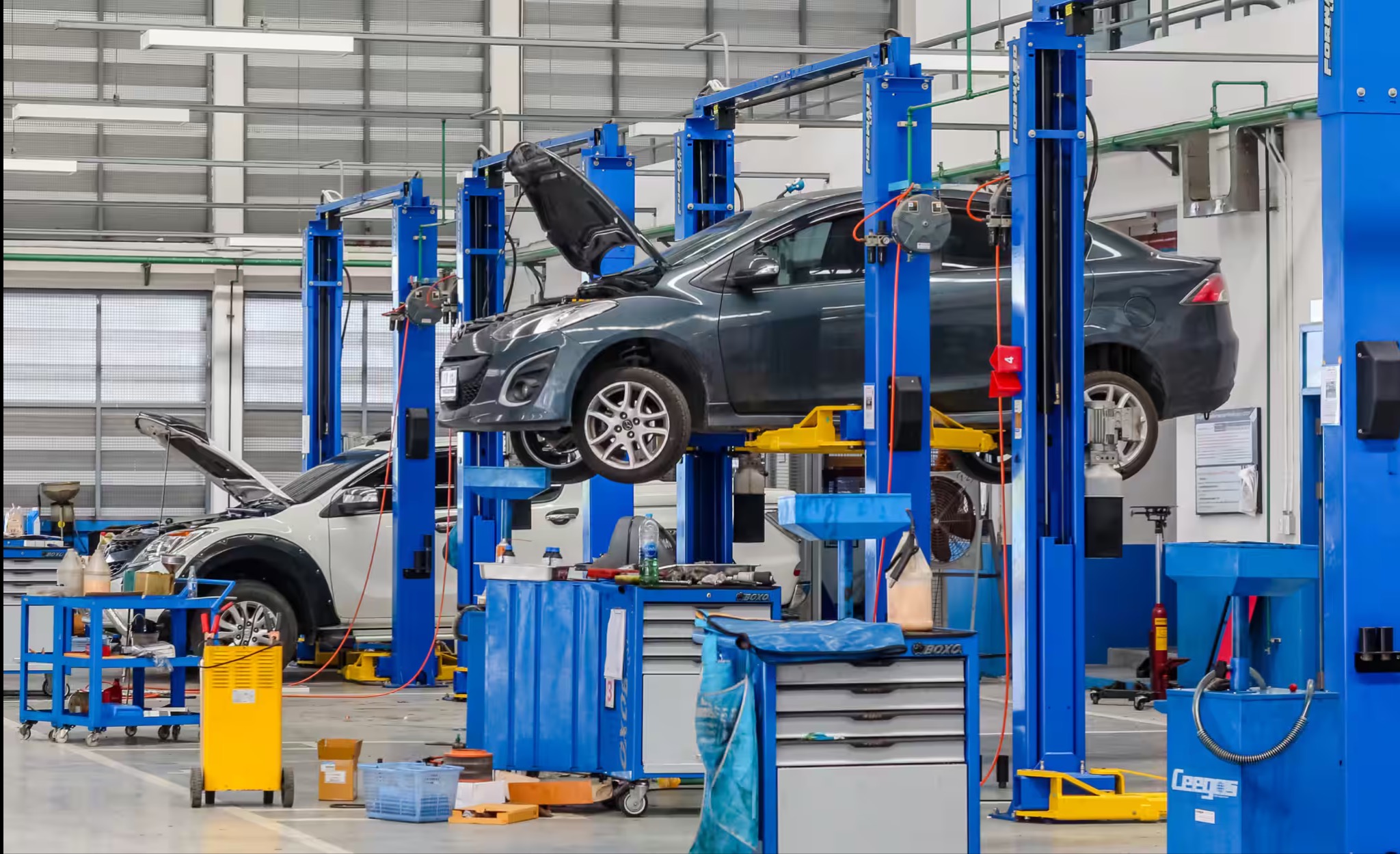 Szary samochód na podnośniku w warsztacie, z otwartą maską. W tle inne podnośniki i narzędzia. Oświetlenie sufitowe, posadzka betonowa.