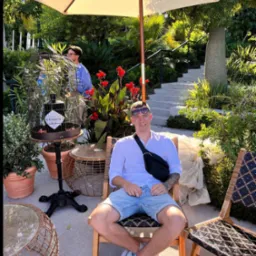 Mężczyzna w błękitnych szortach i białej koszuli siedzi na krześle w ogrodzie, z parasolem nad głową. W tle rośliny i schody.