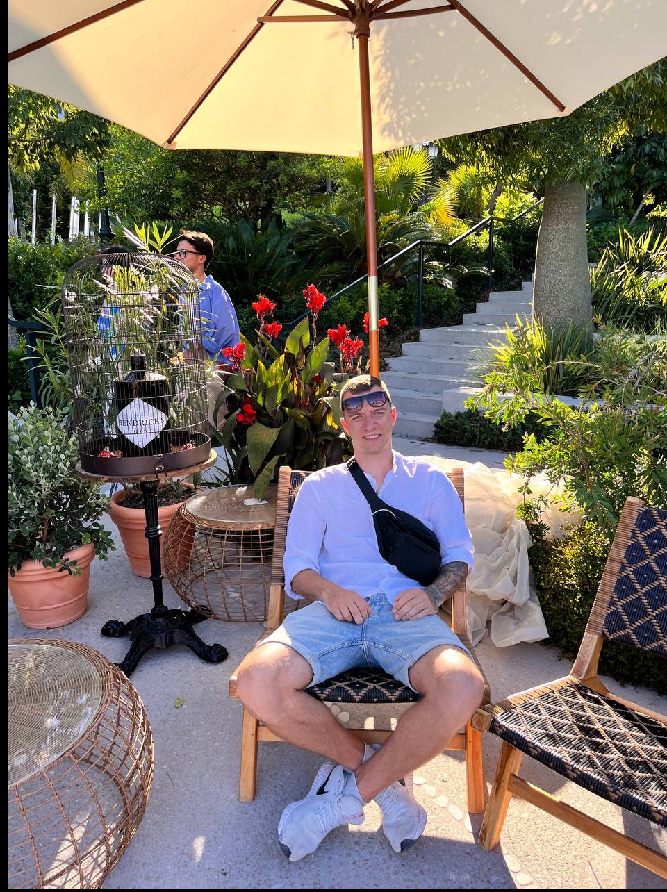 Mężczyzna w błękitnych szortach i białej koszuli siedzi na krześle w ogrodzie, z parasolem nad głową. W tle rośliny i schody.