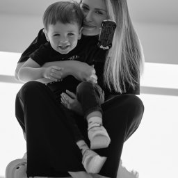 Arina Ihnatenko - Czarno-białe zdjęcie uśmiechniętej kobiety z długimi włosami, trzymającej na kolanach małego chłopca z zabawką w ręku, siedzącej na jasnym tle.
