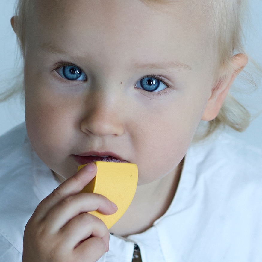 Bliski portret dziecka z jasnymi włosami i niebieskimi oczami, trzymającego w ustach żółty, drewniany element zabawki na neutralnym tle.