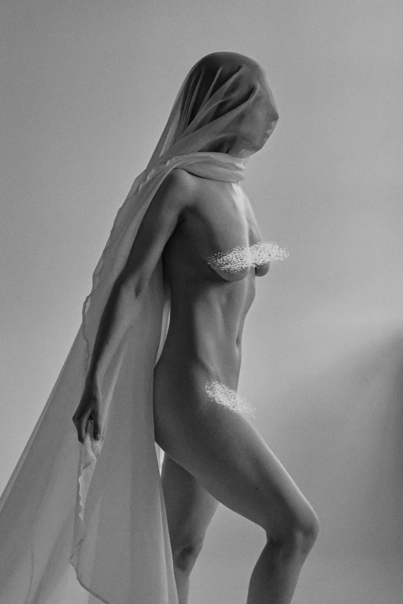 Monochromatyczne ujęcie kobiety owiniętej w prześwitującą tkaninę, z zasłoniętą twarzą i strategicznie ukrytymi fragmentami ciała. Delikatne światło podkreśla teksturę materiału.