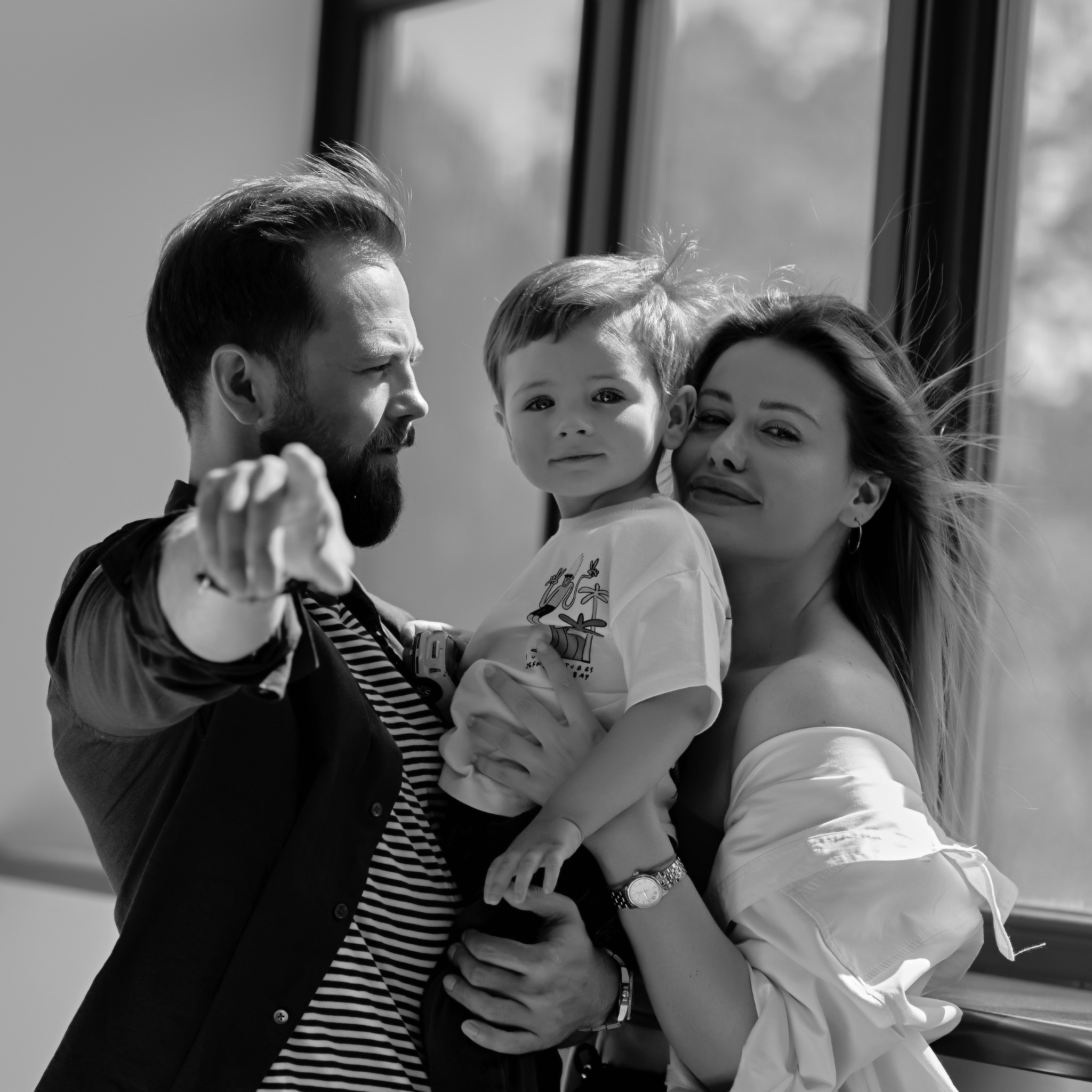 Czarno-białe zdjęcie rodziny: ojciec z brodą wskazuje palcem, matka obejmuje syna. W tle okno z widokiem na zieleń. Ujęcie w naturalnym świetle.