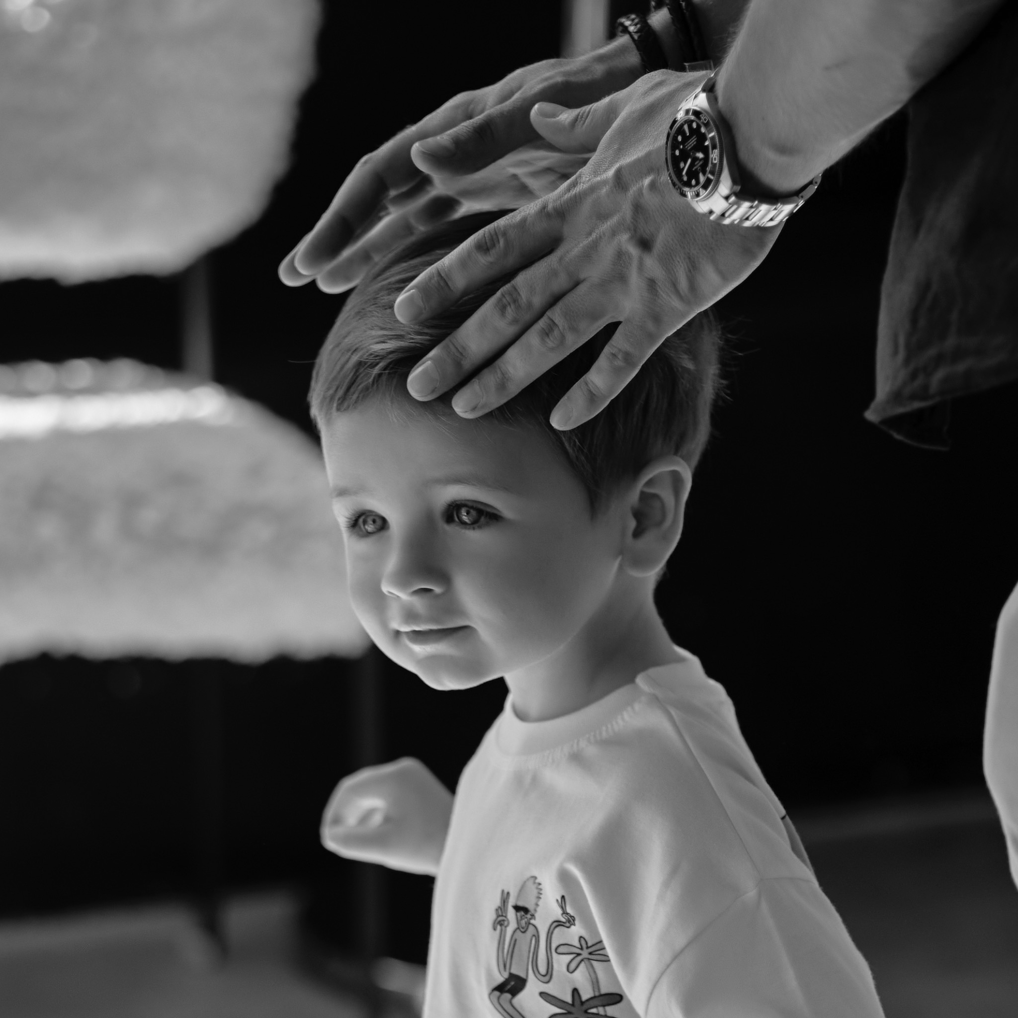 Czarno-białe zdjęcie chłopca z rękami dorosłego na głowie, który ma na sobie zegarek. Ujęcie z bliska, skupione na twarzy dziecka i dłoniach dorosłego.