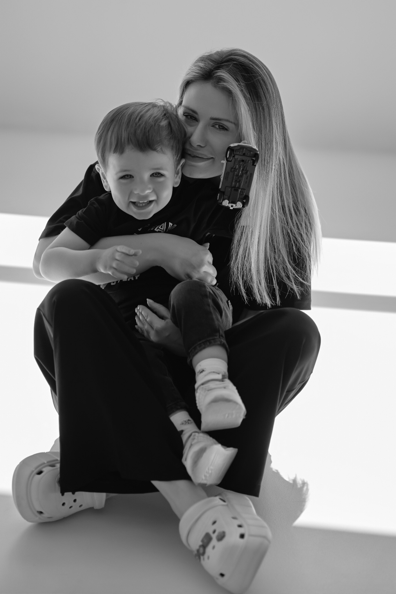 Czarno-białe zdjęcie uśmiechniętej kobiety z długimi włosami, trzymającej na kolanach małego chłopca z zabawką w ręku, siedzącej na jasnym tle.