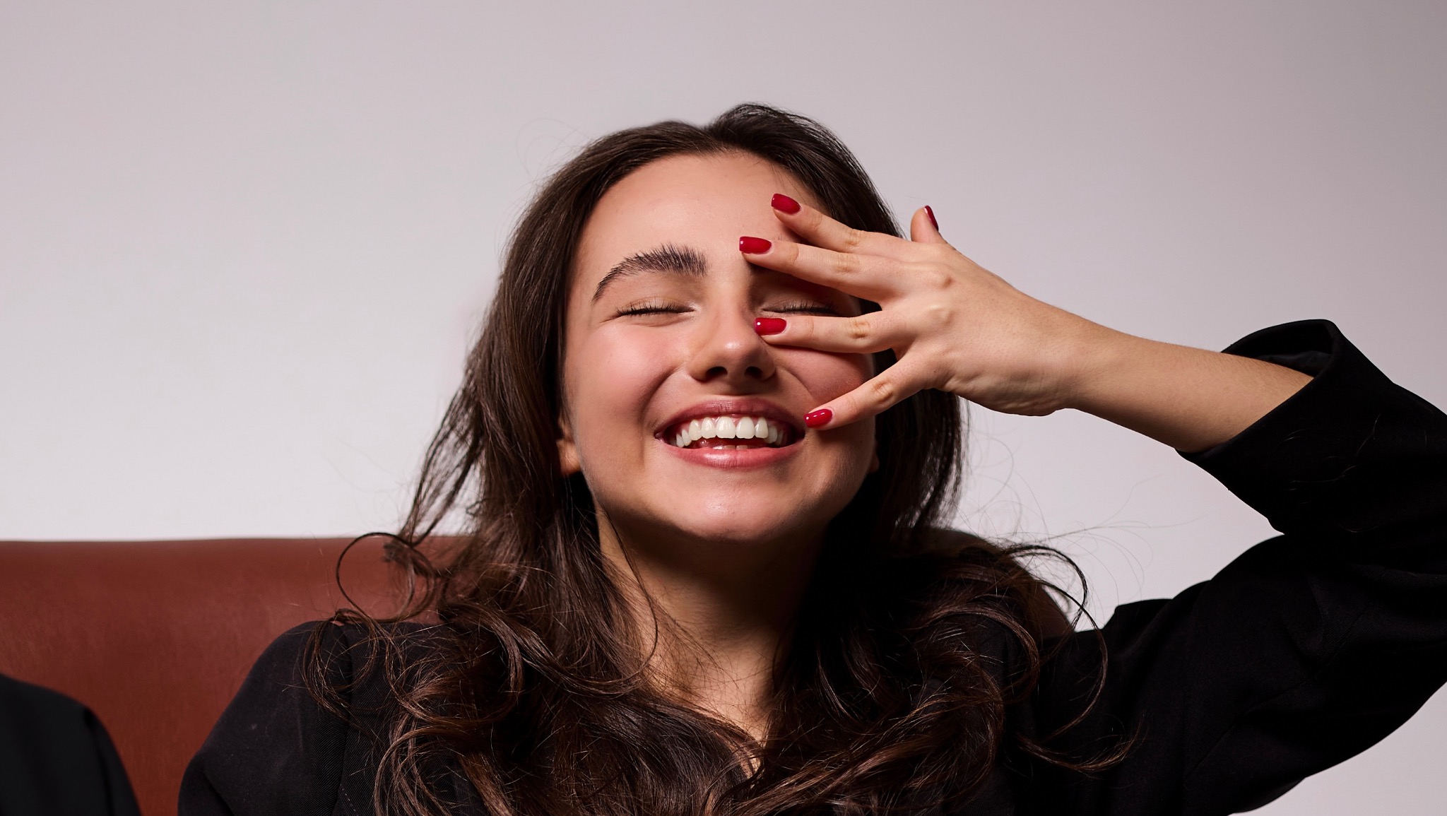 Uśmiechnięta brunetka z czerwonym manicure zakrywa jedno oko dłonią. Jasne tło, ciemna marynarka, brązowa kanapa. Widoczne białe zęby i zadbane brwi.