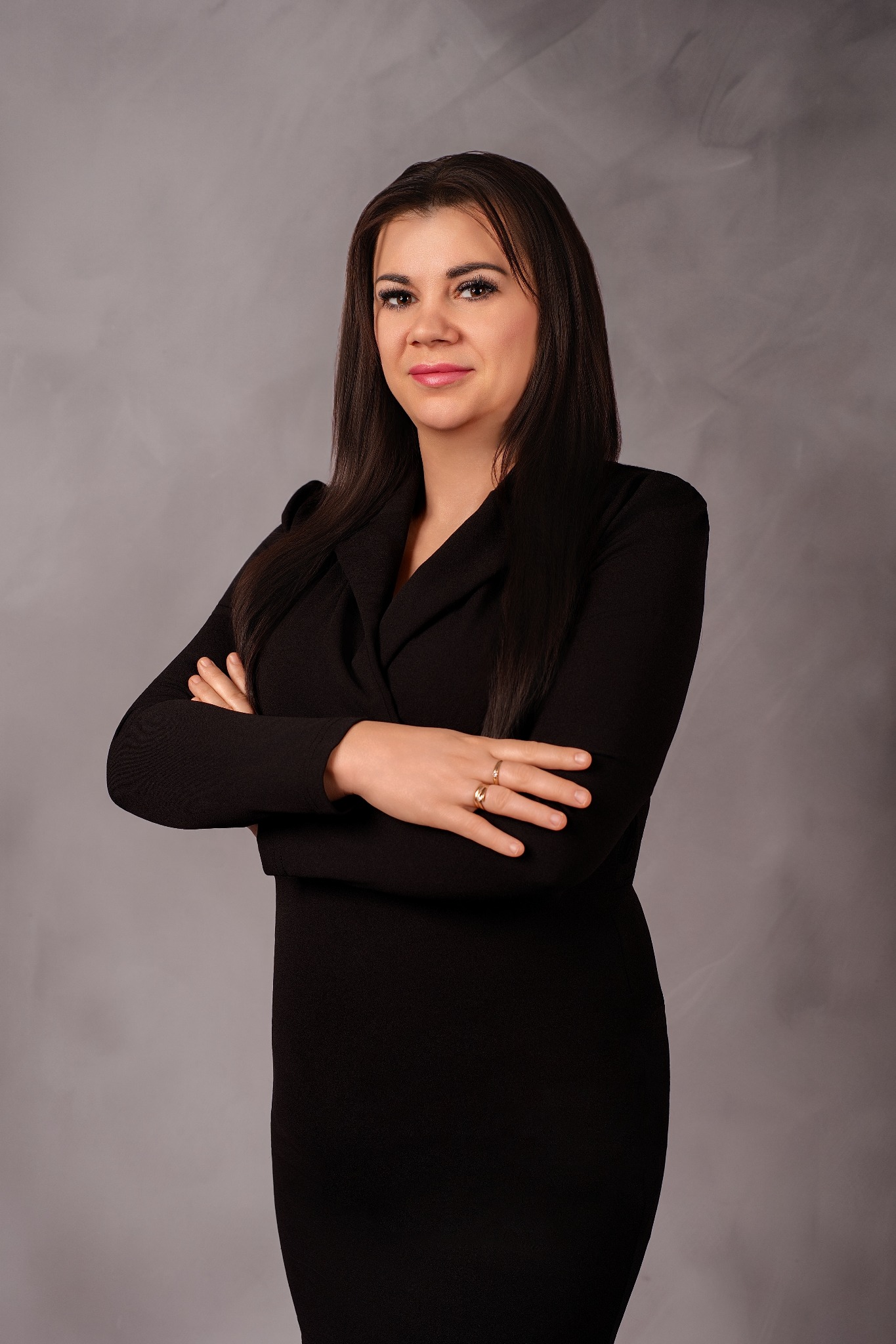 Portret kobiety w czarnej sukience, z założonymi rękami na tle szarego studia. Uśmiechnięta, z pierścionkami na palcach. Profesjonalny wizerunek.