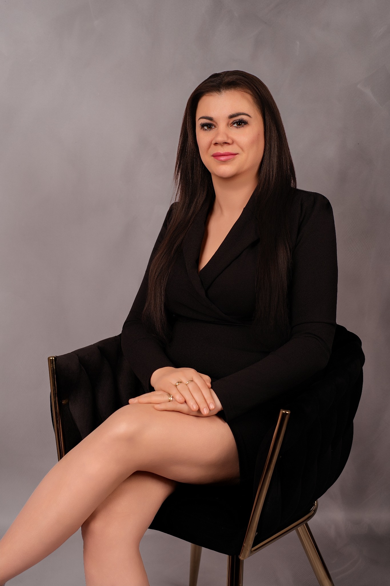 Portret kobiety w czarnej sukience siedzącej na krześle z złotymi nogami na tle szarej ściany. Dłonie splecione na kolanach, uśmiech na twarzy.