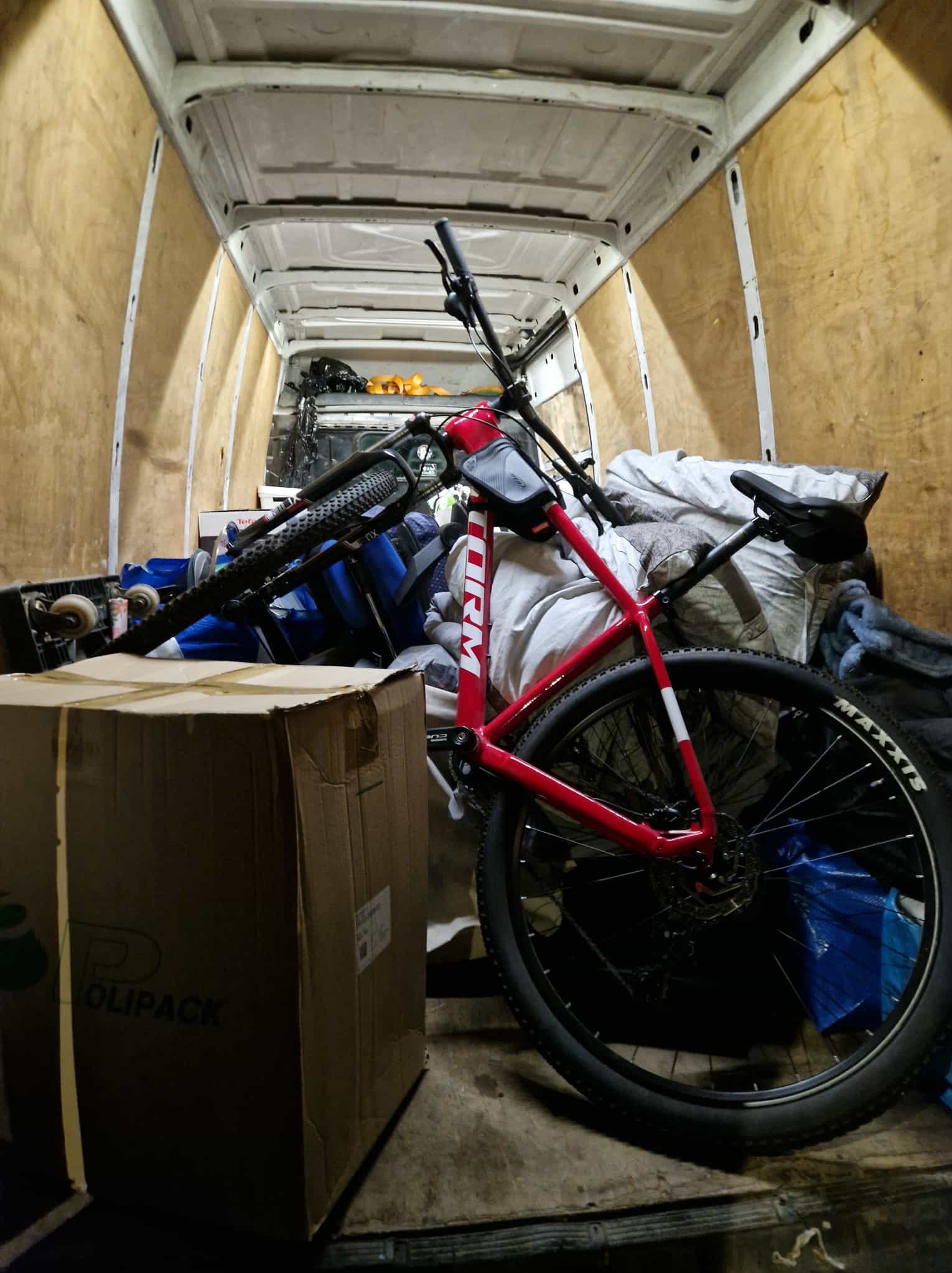 Wnętrze furgonetki z rowerem górskim, kartonem i zabezpieczeniami. Widoczne drewniane panele boczne i biały sufit pojazdu. Rower oparty o karton, gotowy do transportu.