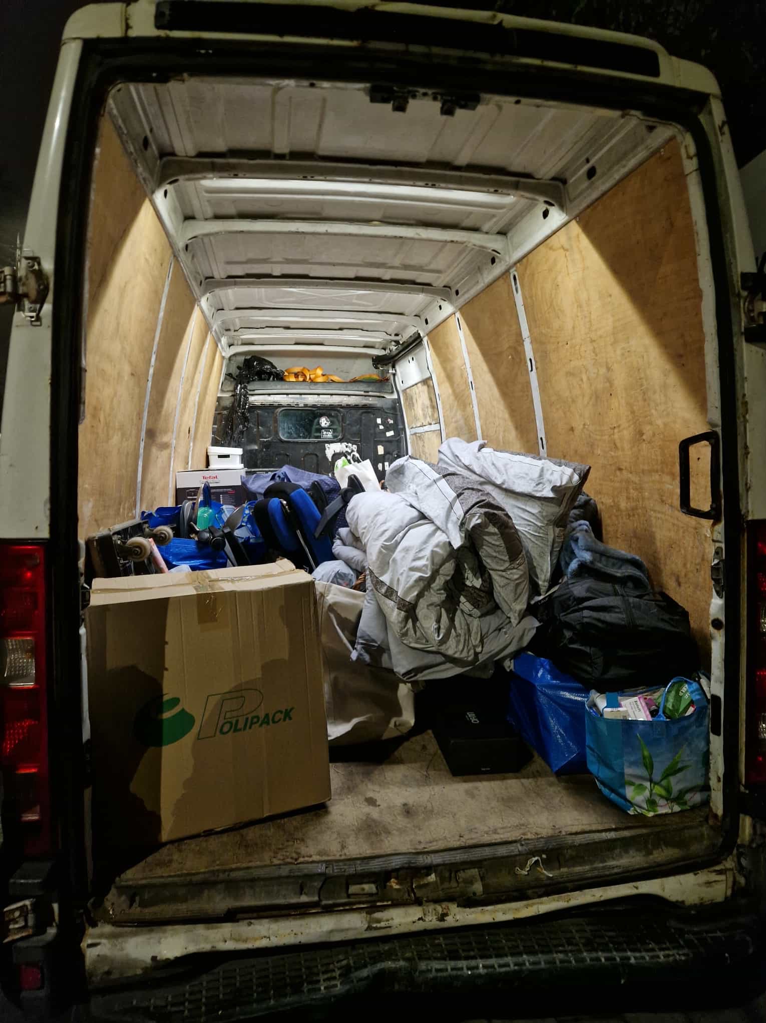 Wnętrze białego busa załadowane kartonami, torbami i workami, przygotowane do transportu. Widoczne zabezpieczenia ścian i podłogi. Nierównomierne oświetlenie.