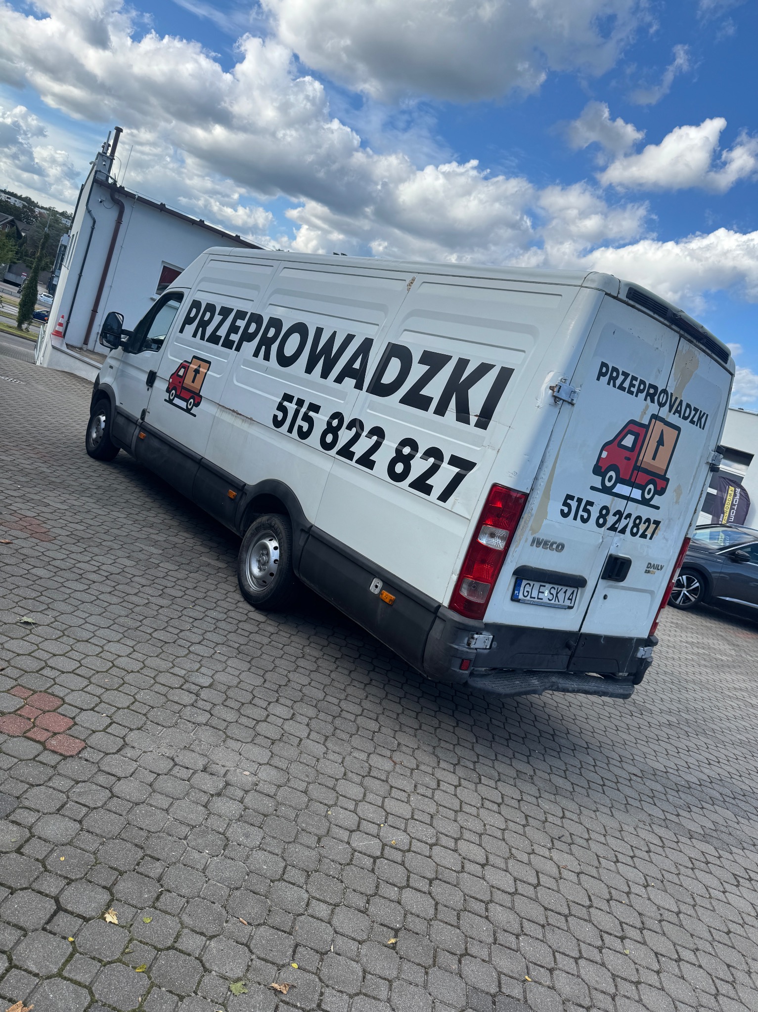 Biały bus Iveco z napisem 'Przeprowadzki' i numerem telefonu, zaparkowany na kostce brukowej. Widoczne logo firmy z pudełkiem na pace. Błękitne niebo z chmurami w tle.